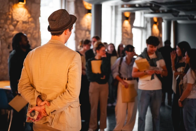Guillaume Barray, alias Gentledice, face au public. Il est au premier plan de la photo, une carte caché dans le dos. Le public est à l'arrière plan de la photographie, observant le maître du jeu.