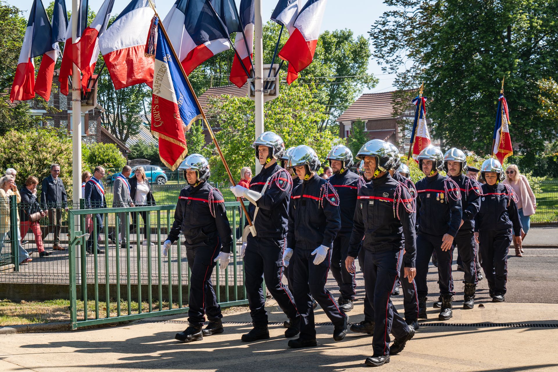 Les pompiers de Grand Quevilly défilent en formation avec des drapeaux. Ils sont une vingtaine et portent l'uniforme des sapeurs pompiers.